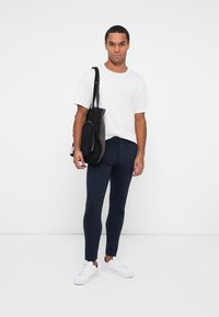 Pantaloni slim-fit blu navy, t-shirt in cotone bianca, indossati con una borsa scura. Il modello è in piedi contro uno sfondo bianco semplice, indossando sneakers bianche.