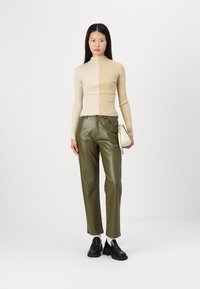 Bežani rebrasti turtleneck, maslinasto zeleni hlače od umjetne kože i crne mokasine. Model drži malu bijelu torbicu, u pozadini bijela pozadina.