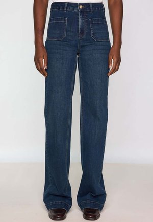 Person iført mørkeblå jeans med høj talje, forlommer med lapper og brune sko står mod en ensfarvet baggrund.