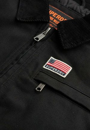 Zwarte jas met een corduroy kraag, zilveren ritsuitstekers en een geborduurd Amerikaanse vlag patch met de tekst "SUPERDRY" eronder.