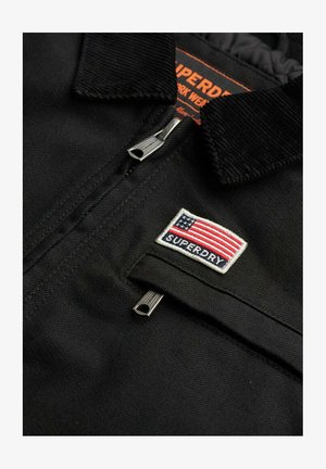 Zwarte jas met een corduroy kraag, zilveren ritsuitstekers en een geborduurd Amerikaanse vlag patch met de tekst "SUPERDRY" eronder.