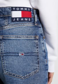 Detailní záběr zadní kapsy modrých džín s nášivkou značky Tommy Jeans a malou vyšívanou vlajkou, nošeno s bílým topem.