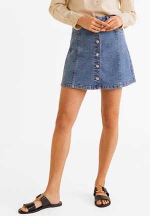 Mujer con minifalda vaquera abotonada al frente, camiseta beige de manga larga y sandalias negras tipo slide, de pie con las manos cerca de la cintura.