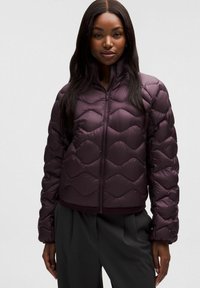 Kobieta w ciemnofioletowej pikowanej kurtce z wysokim kołnierzem, zamkiem błyskawicznym z przodu i długimi rękawami, w połączeniu z luźnymi czarnymi spodniami.