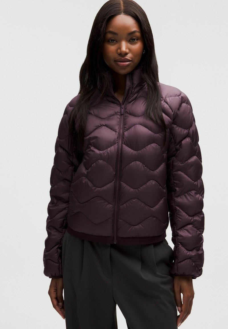 Kobieta w ciemnofioletowej pikowanej kurtce z wysokim kołnierzem, zamkiem błyskawicznym z przodu i długimi rękawami, w połączeniu z luźnymi czarnymi spodniami.