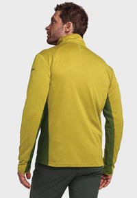 Langarmjacke, gelbe Vorderseite mit grünen Seitenpaneelen, strukturierten Stoff, hoher Kragen, taillierter Schnitt, mit subtiler Logo-Applikation am Ärmel.