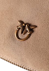Beige Wildleder-Handtasche mit einem markanten bronzefarbenen Hardware-Design, das zwei Vögel und einen runden Ring umfasst, ergänzt durch eine genähte Kante.