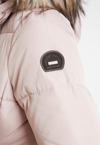 Světle růžová prošívaná bunda s hladkou texturou, zdobená hnědým kulatým štítkem s nápisem "OUR STORY SINCE 1995" a stříbrnými detaily.
