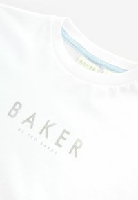 Bijela pamučna majica s srebrnim logotipom "BAKER" i tekstom "by TED BAKER". Okrugli izrez i glatka tekstura. Oznake su vidljive unutra.