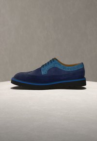 Chaussure Oxford en daim bleu pour homme avec détails en aile et semelle noire avec accent bleu, présentée sur une surface ronde et claire.