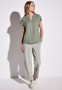 Blusa verde chiaro a maniche corte con scollo a V, abbinata a pantaloni larghi grigi chiari. Il modello indossa sneakers bianche; texture morbida e vestibilità rilassata.