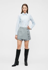 Γυναίκα που στέκεται με το χέρι στη μέση, φορώντας ανοιχτό μπλε πουκάμισο με κουμπιά, γκρι ασύμμετρο σορτς-φούστα και μαύρες μπότες μέχρι το γόνατο σε λευκό φόντο.