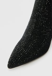 Μαύρο τακούνι με αιχμηρή μύτη, φτιαγμένο από υφασμάτινη υφή διακοσμημένο με μικρές παγιέτες, δημιουργώντας ένα λαμπερό αποτέλεσμα στην επιφάνεια.