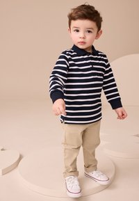 Pruhované polokošeľa v námorníckej modrej a bielej farbe, béžové nohavice a biele tenisky s červenými doplnkami. Dieťa stojí na kruhovej platforme.