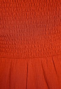 Texturerad orange tyg med samlat yta, med en smockad byst och utsvängd kjol. Tyget verkar mjukt och något elastiskt.