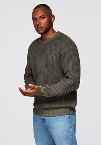 Suéter de punto gris-verde con patrón texturado, puños y dobladillo acanalados, combinado con jeans de mezclilla azul claro. El modelo posa con las manos abiertas.