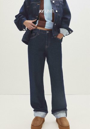 Dark blue denim jacket and matching jeans with cuffed hems. Underneath is a color-blocked brown and blue sweatshirt. Brown footwear completes the outfit.