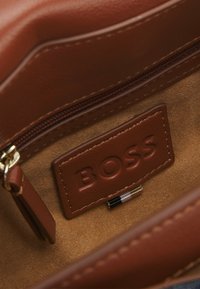 Braunes Lederinterieur einer Tasche mit einer Reißverschlusstasche und einem rechteckigen Markenschild, das mit "BOSS" in einem dunkleren Braun geprägt ist.