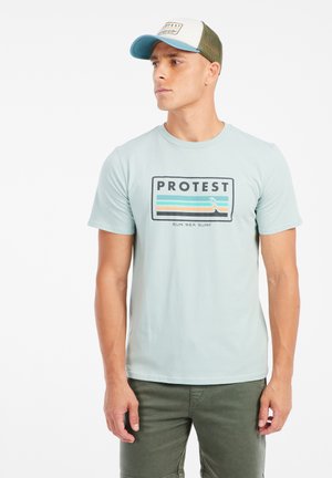 Hellblaues Baumwoll-T-Shirt mit schwarzem und farbigem Paneeldesign, das das Wort "PROTEST" und Wellen-Grafiken zeigt. Kombiniert mit olivgrünen Shorts und einer Kappe.