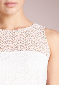 Vit klänning med en texturerad spetsöverdel med ett blommönster, som kontrasterar med en slät nedre del, ärmlös design och rund halsringning.