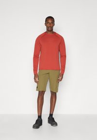 Rød langermet skjorte med teksturert stoff, kombinert med olivengrønne shorts. Personen har på seg svarte joggesko med mørke sokker.