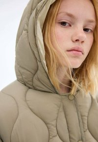 Beige quiltad pufferjacka med huva, med upphöjda sömmar och knappstängning. Mjuk textur och slät innertyg.