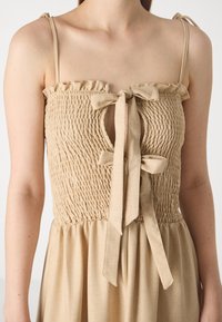 Beige jurk met smock-bovenstuk, gerimpelde halslijn en strikdetails aan de voorkant. Verstelbare banden, vloeiende rok en een zachte stofstructuur.