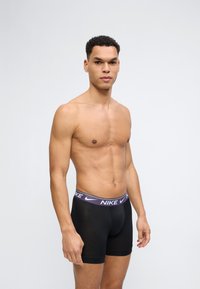 Ευτραφής άνδρας με κοντά, σγουρά μαλλιά, φοράει μαύρα σλιπ της Nike με μωβ ζώνη μέσης, στέκεται μπροστά σε απλό φωτεινό φόντο.