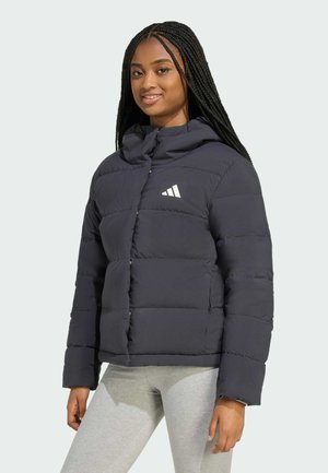 Młoda kobieta z długimi warkoczami, ubrana w czarną kurtkę puchową z białym logo i jasnoszare legginsy, stoi na tle jednolitego tła.