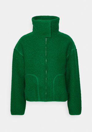 Person wearing green textured hooded jacket over black and white plaid shirt, patchwork jeans, and white sneakers, hands in pockets.