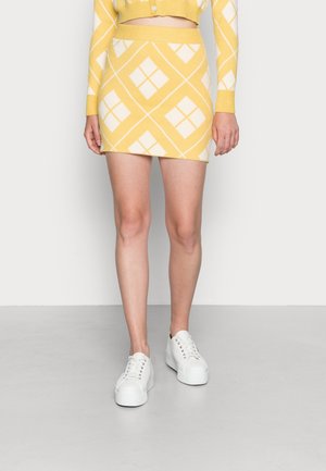 Femme portant une mini-jupe jaune et blanche à motifs assortie à un pull court et des baskets blanches à plateforme, debout sur un sol gris.
