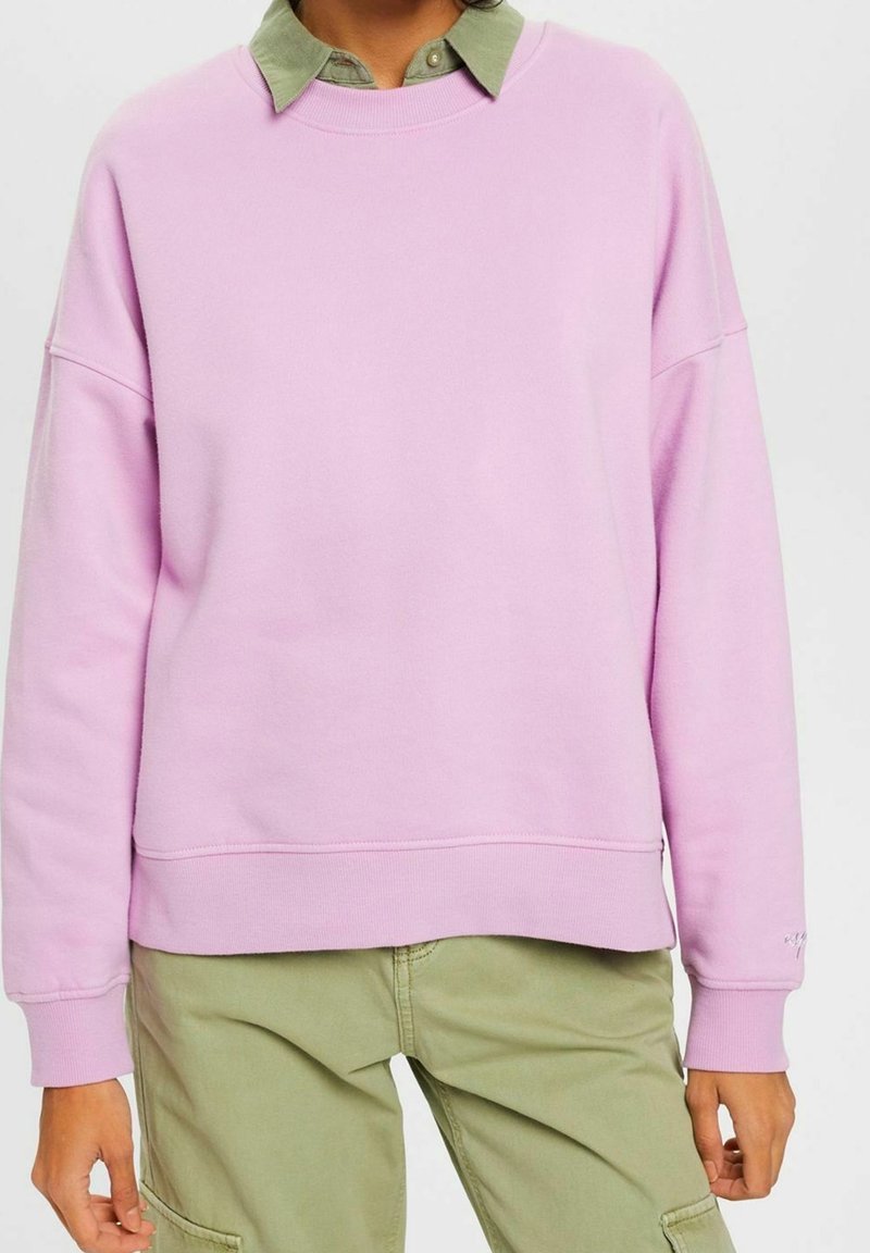 Sweatshirt rose clair avec un col rond, des épaules dénudées et des poignets côtelés, superposé à une chemise verte à col. Texture douce.