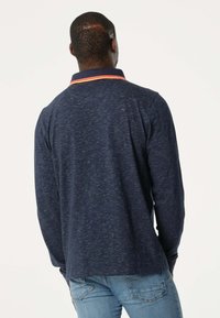 Marineblauwe longsleeve poloshirt met een contrasterende oranje kraag. Zachte textuur, ontspannen pasvorm en zichtbare stikseldetails. Draagt met lichtblauwe spijkerbroek.