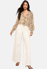 Blusa beige con volant e stampa zebraprim, annodata al colletto, abbinata a pantaloni bianchi a gamba larga e sandali neri con tacco su uno sfondo neutro.