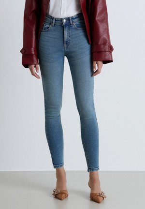 Donna con jeans skinny azzurri chiaro, tacchi marroni a punta con fibbie, camicia bianca e giacca in pelle rosso scuro, in piedi su pavimento grigio.