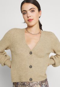 Beige gebreide cardigan met een V-hals, geribbeld dessin en vijf donkere knopen aan de voorkant. De mouwen zijn iets pof.