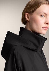Zwarte capuchonjas met een hoge kraag en rits. Gladde textuur, slank ontwerp, minimale naden en een eigentijdse stijl.
