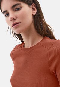 Mujer joven con ojos azules y cabello castaño lleva una blusa de canalé color óxido frente a un fondo blanco liso.