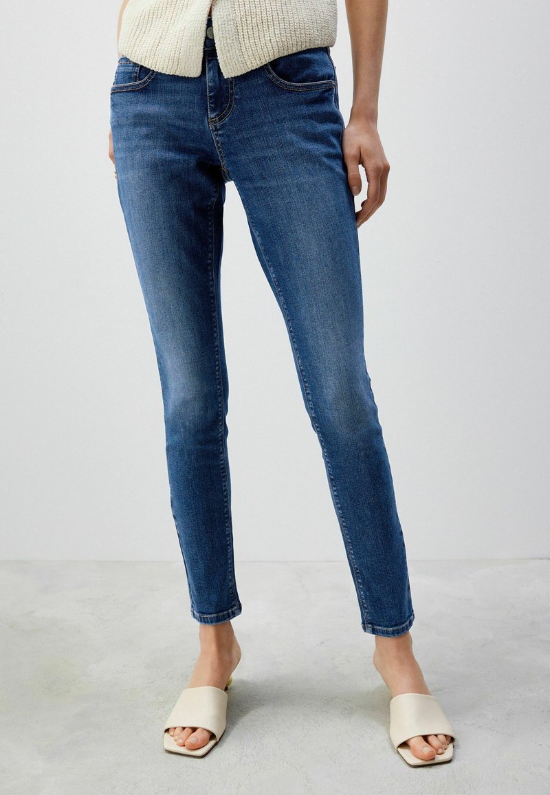 Blå denim skinny jeans med en medelhög midja, sydda detaljer och en slät textur, i kombination med krämfärgade slidessandaler.