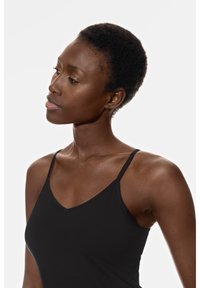 Zwarte mouwloze top met dunne schouderbandjes, met een V-hals ontwerp en een gladde, gestructureerde stof. Model toont een neutrale pose.