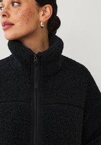 Zwarte fleecejack met een hoge kraag, ritssluiting en textuurstof. Voorzien van een contrasterende, gladde rits en stikdetails.