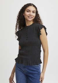 Blouse noire texturée avec un col montant, des manches courtes à volants et un détail de bouton sur le devant, dotée d'une ourlet évasé.