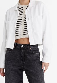 Vrouw in een witte geknipte button-up jacket over een gestreept crop top en hoge zwarte jeans, staande tegen een witte achtergrond.