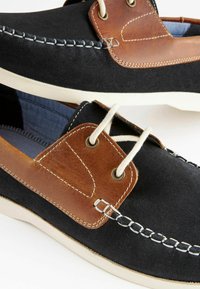 Scarpe da barca con tomaia in suede nero, dettagli in pelle marrone, lacci crema e suole in gomma bianche. Cuciture decorative lungo i bordi.