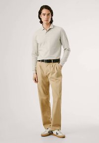 Šviesiai pilkas ilgomis rankovėmis polo marškinėlis, smėlio spalvos raukšlėtos kelnės, juodas diržas ir balti sportbačiai su juodais akcentais. Paprastas, laisvo kirpimo dizainas.