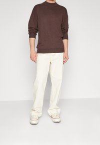 Brązowy sweter z dzianiny o wysokim kołnierzu, w połączeniu z luźnymi kremowymi spodniami i jasnymi sneakersami, tworząc casualowy, warstwowy wygląd.