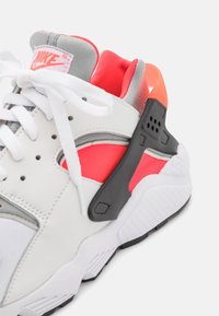 Sneakers Nike Air Huarache z białej skóry, szarymi akcentami, czarnym wsparciem pięty z gumy, żywymi czerwonymi i pomarańczowymi detalami oraz teksturowaną podeszwą.