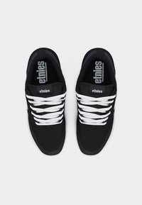 Ζευγάρι μαύρα αθλητικά παπούτσια Etnies με λευκά κορδόνια, εμφανιζόμενα από ψηλά σε απλό φωτεινό φόντο.