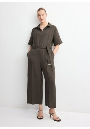 Mujer vistiendo un mono con cinturón en color oliva oscuro, de manga corta y pantalones de pierna ancha, combinado con sandalias de tacón negras con tiras.