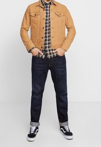 Giacca di denim tan con due tasche sul petto, indossata sopra una camicia a quadri blu navy e gialla, abbinata a jeans scuri e sneakers nere.